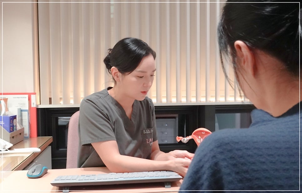 회색 스크럽 여성 스태프가 책상에 앉아 두 손을 모으고 환자(뒷모습, 남색 상의)와 마주보며 이야기하는 모습