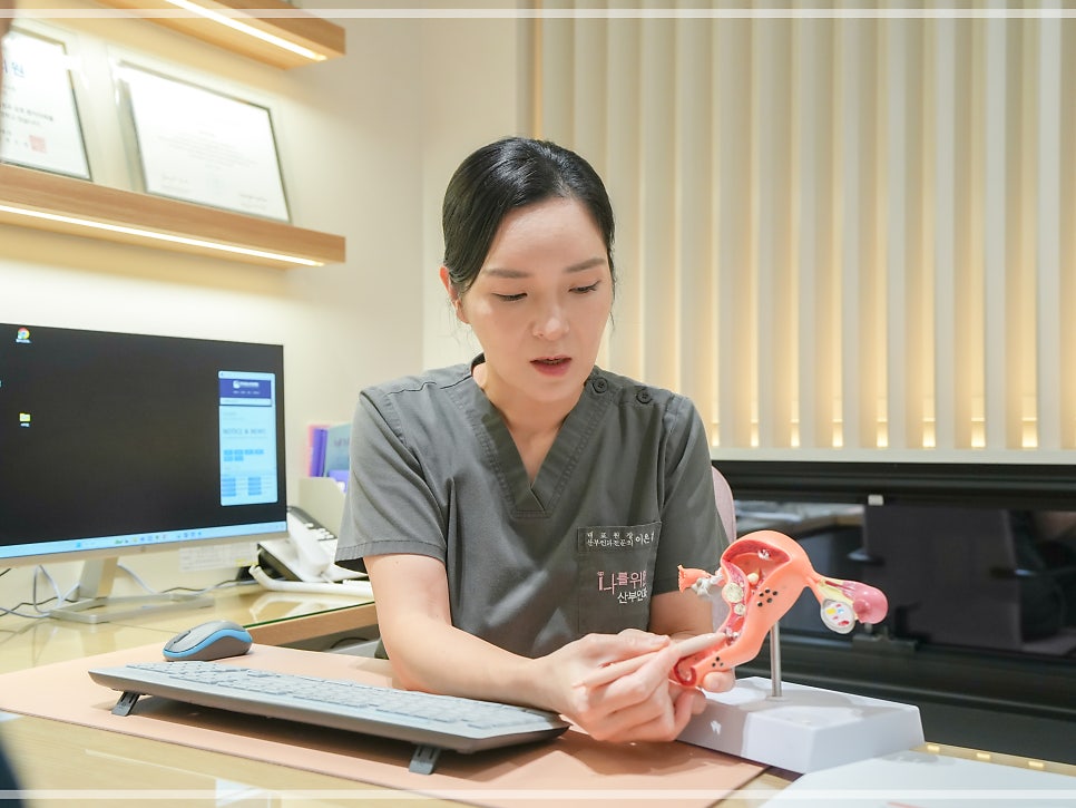 벽 선반에 액자(인증서로 보임)가 걸린 진료실에서 의료진이 분홍색 자궁 모형을 들고 위를 올려다보며 말하고 