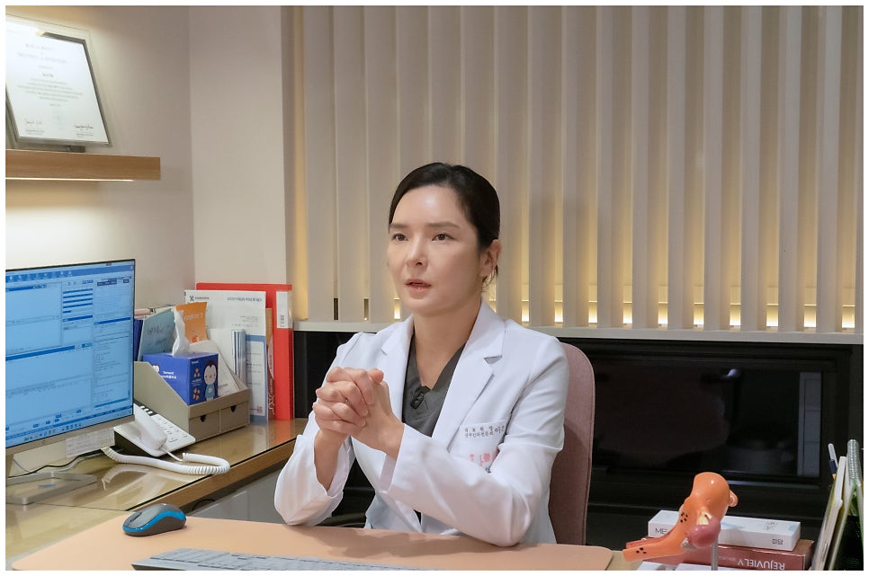 흰 가운 입은 여성 의료진이 책상에 앉아 손을 모으고 말하는 인터뷰형 컷